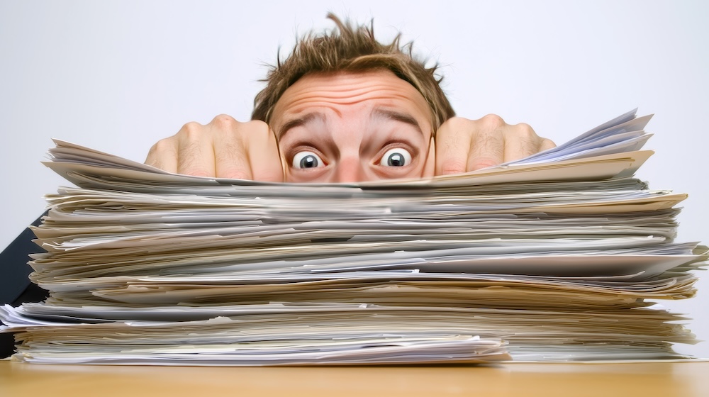 A man expresses frustration as he stares at an overwhelming pile of paperwork on his desk, symbolizing work stress and anxiety in a busy office environment. Concept AI needed to help with busywork.