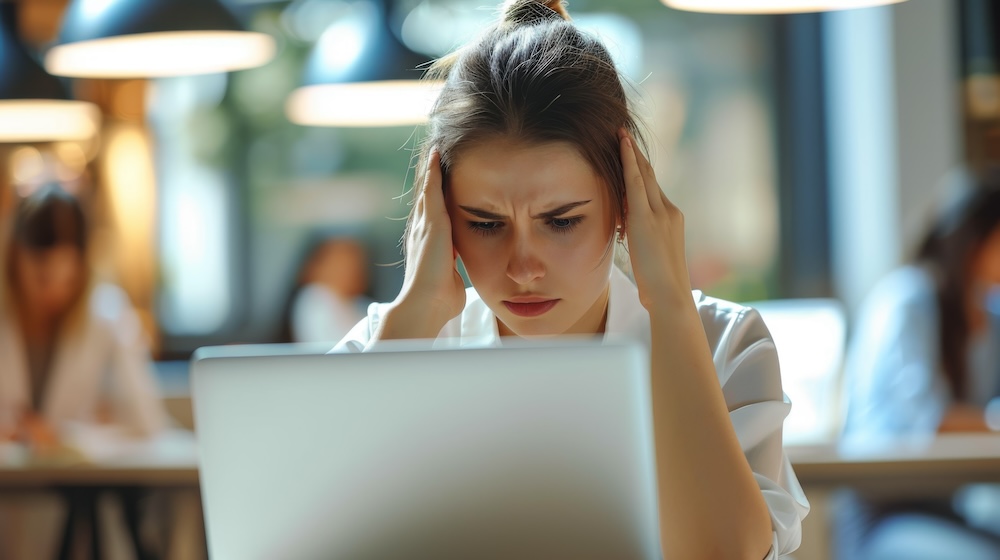 stressed young woman with her hands on her head is looking at a laptop screen with a worried expression, possibly dealing with a problem or challenging work